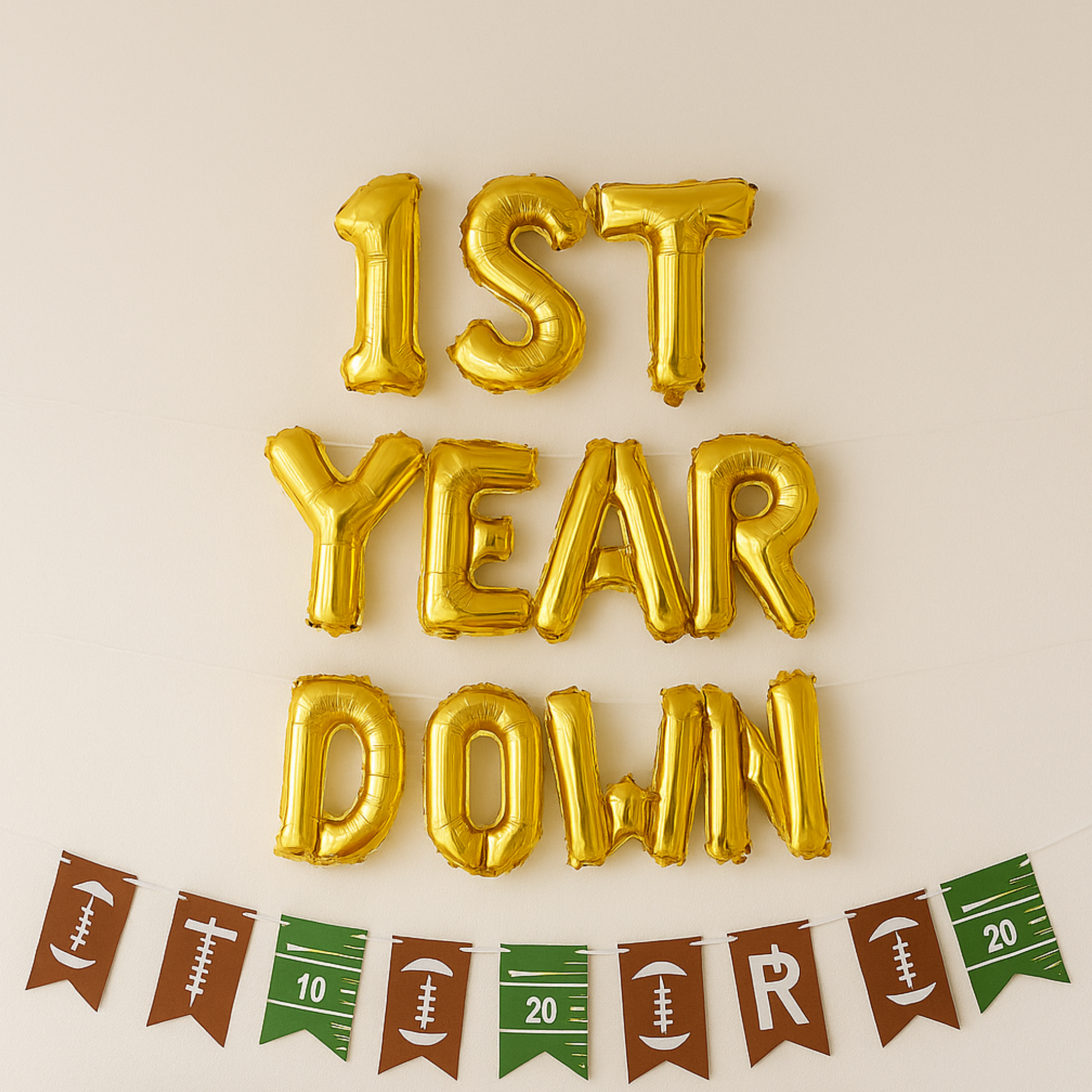Gold foil balloons spelling '1st Year Down' on a beige background with football-themed bunting.