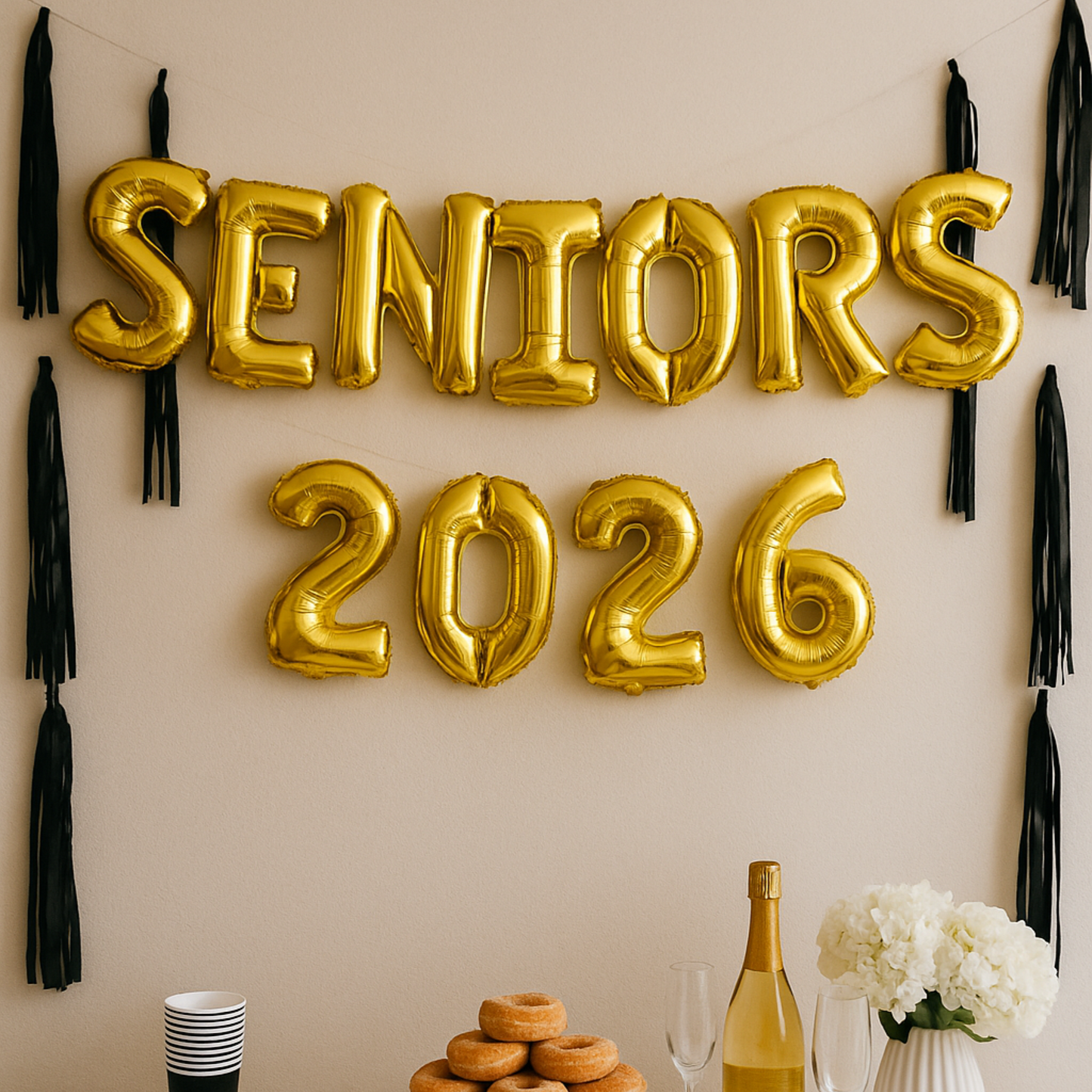 Gold 'SENIORS 2026' balloons on a wall with tassels and a table with a bottle, glasses, and flowers.