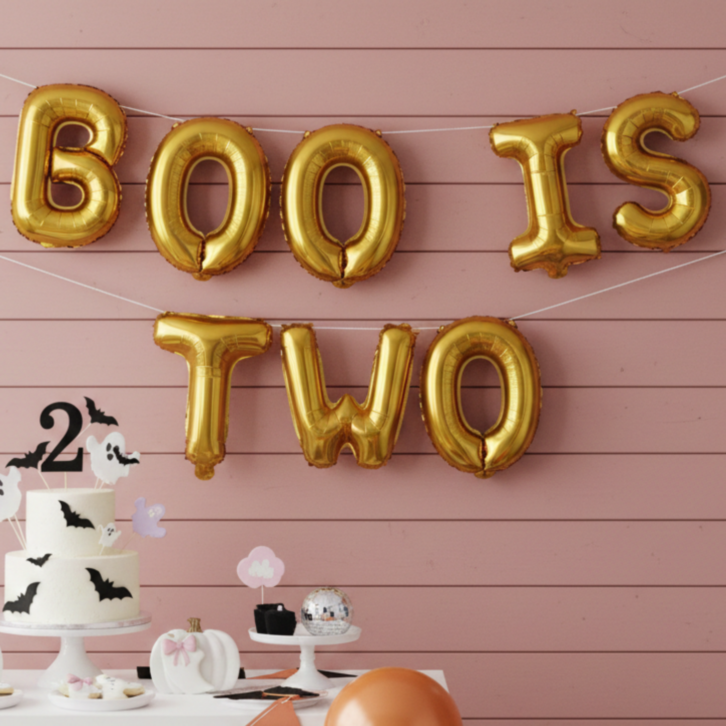 Halloween-themed birthday party setup with gold 'BOO IS TWO' balloons, cake, and balloons on a pink wooden wall.