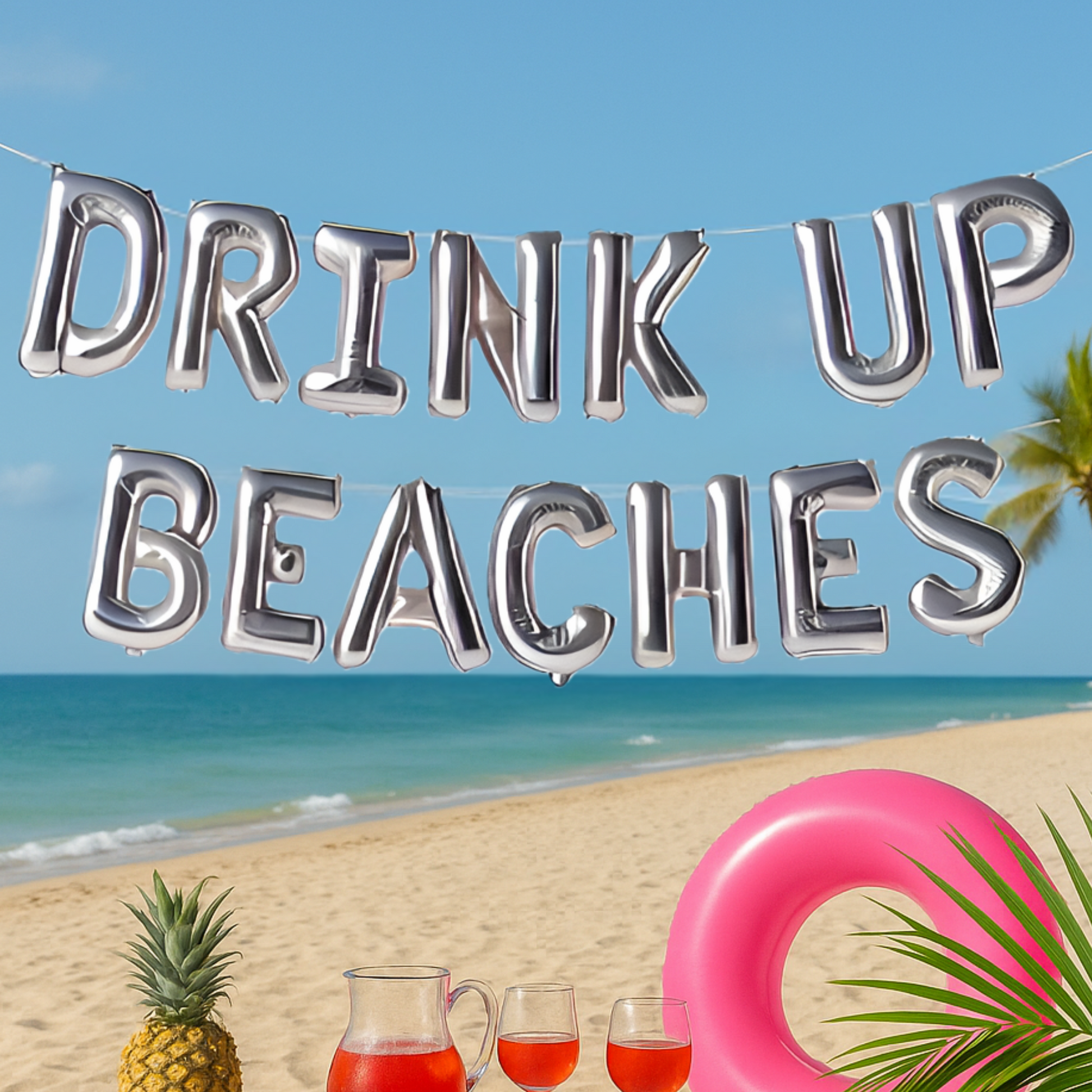 Silver 'DRINK UP BEACHES' balloon banner on a beach with pineapple, drinks, and pink ring.