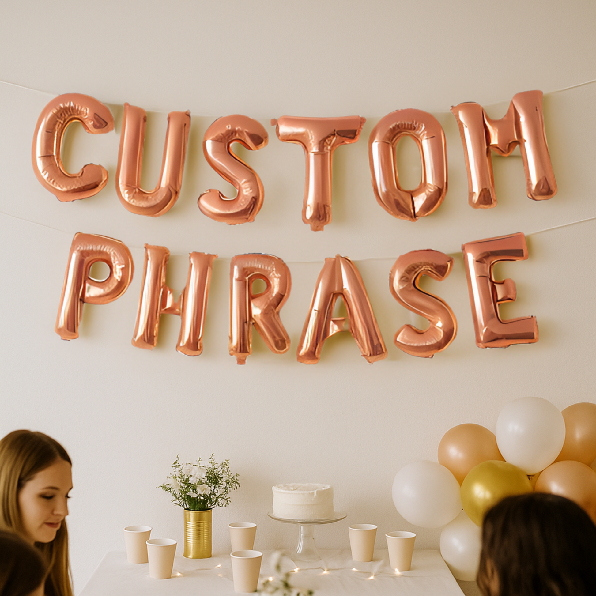 Rose gold letter balloons spelling 'CUSTOM PHRASE' above a table with a cake and balloons.