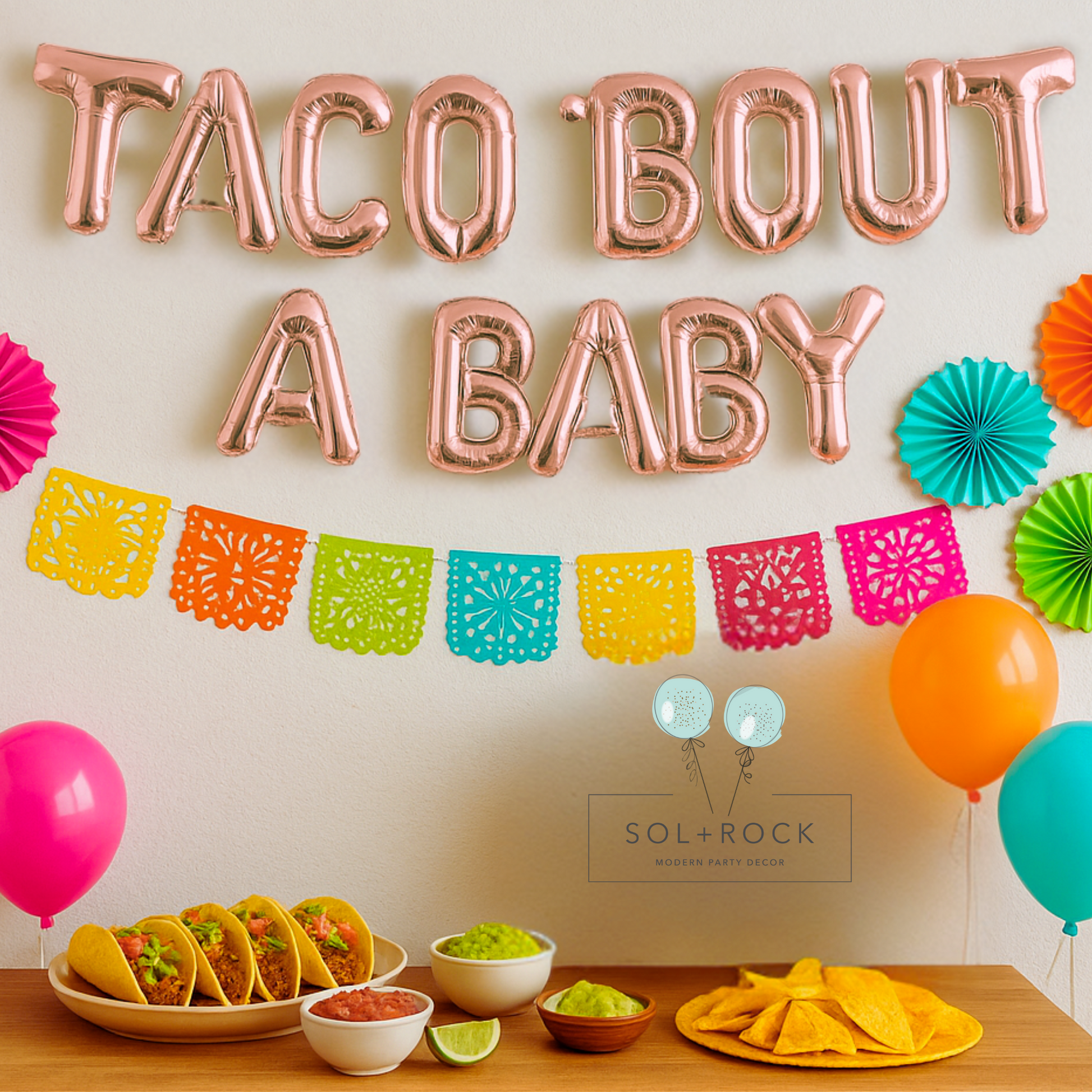 Decorative setup with 'Taco Bout A Baby' balloons, colorful paper banners, and food items on a table.
