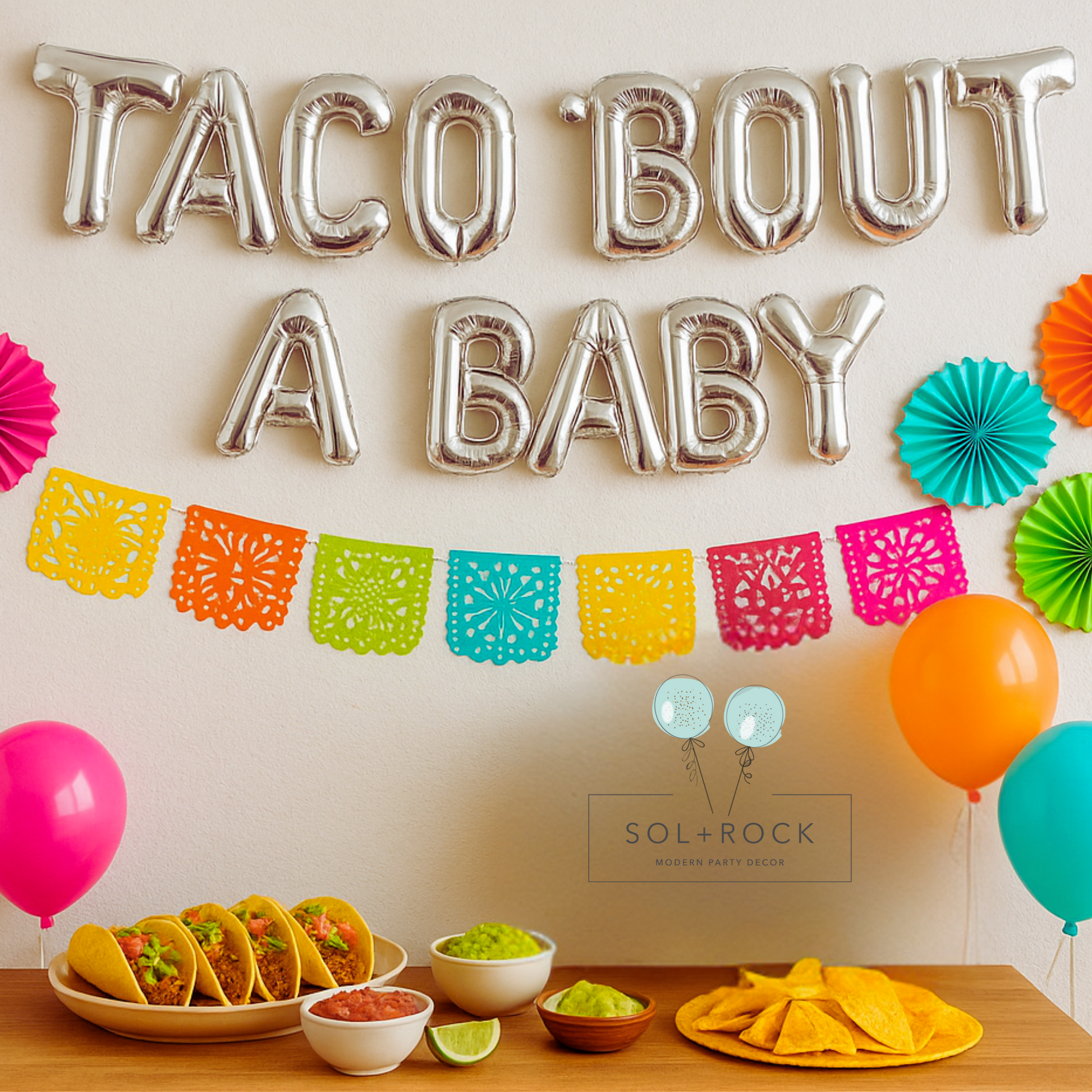 Decorative setup with 'Taco Bout A Baby' balloons, colorful balloons, and party food on a table.