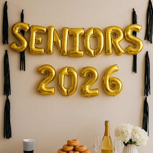 Gold 'SENIORS 2026' balloons on a wall with tassels and a table with a bottle, glasses, and flowers.